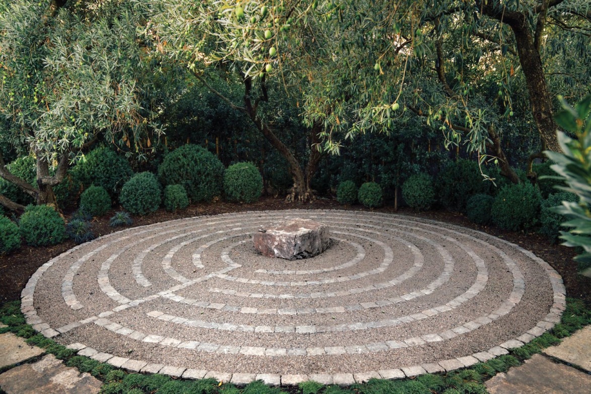 Circular stone labyrinth surrounded by lush greenery and trees in a peaceful garden setting.