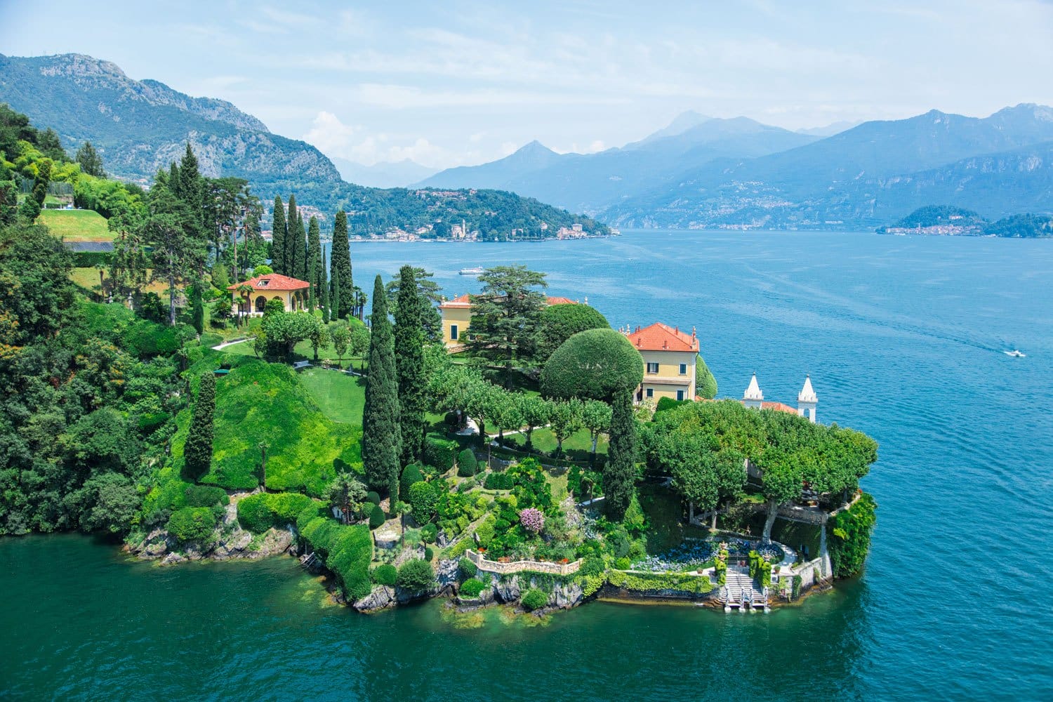 Scenic aerial view of lush greenery, historic architecture, and mountains by a serene blue lake edge.