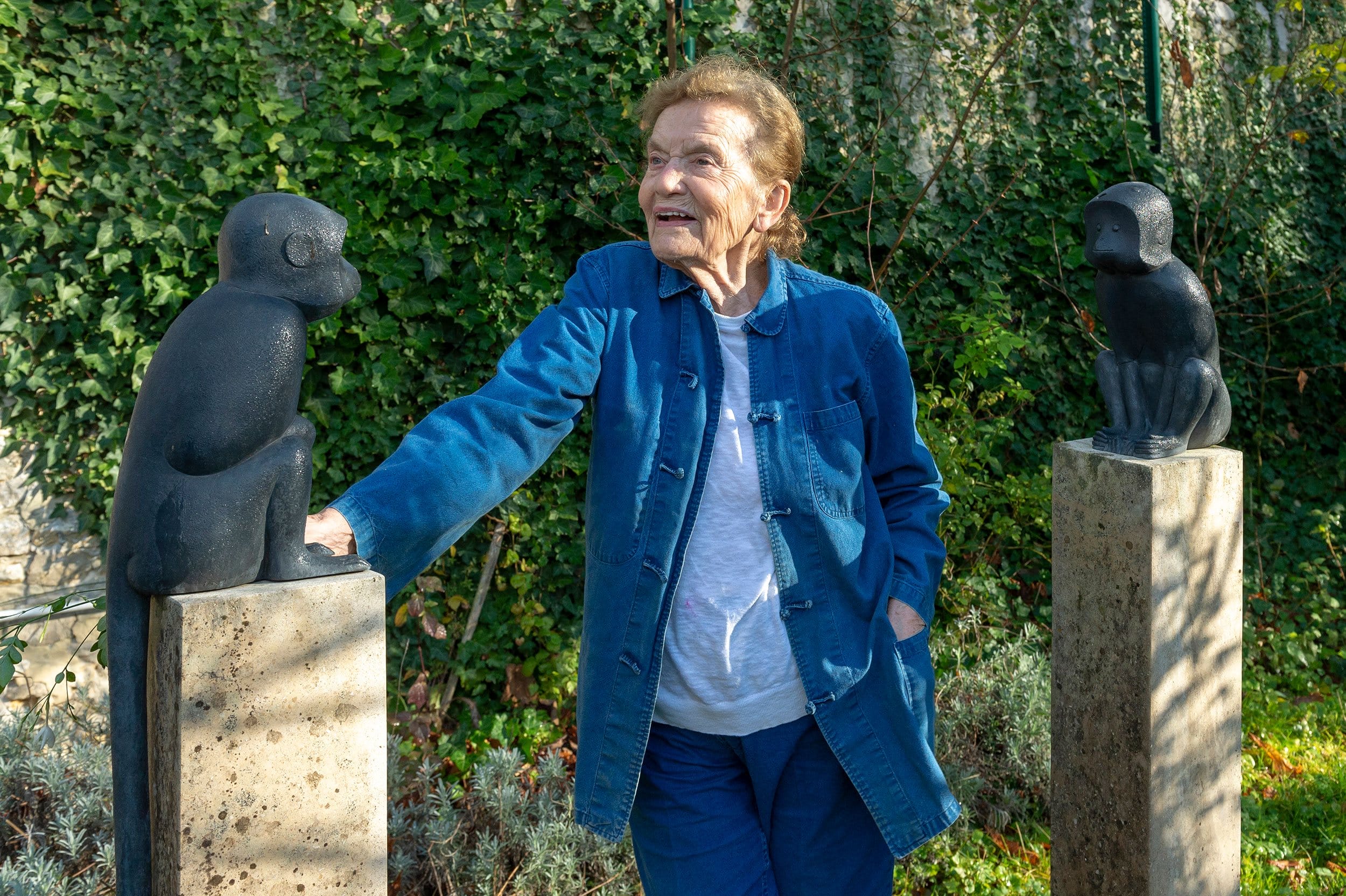 Older woman standing between two monkey sculptures outdoors, wearing a blue jacket, smiling amidst greenery.