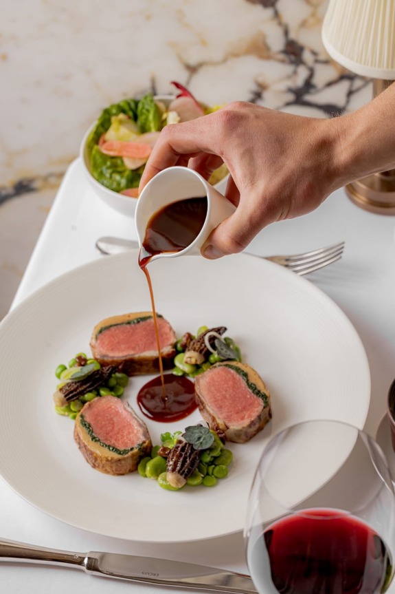 Gourmet dish of sliced meat with a sauce being poured over, accompanied by vegetables and a glass of red wine on the table.