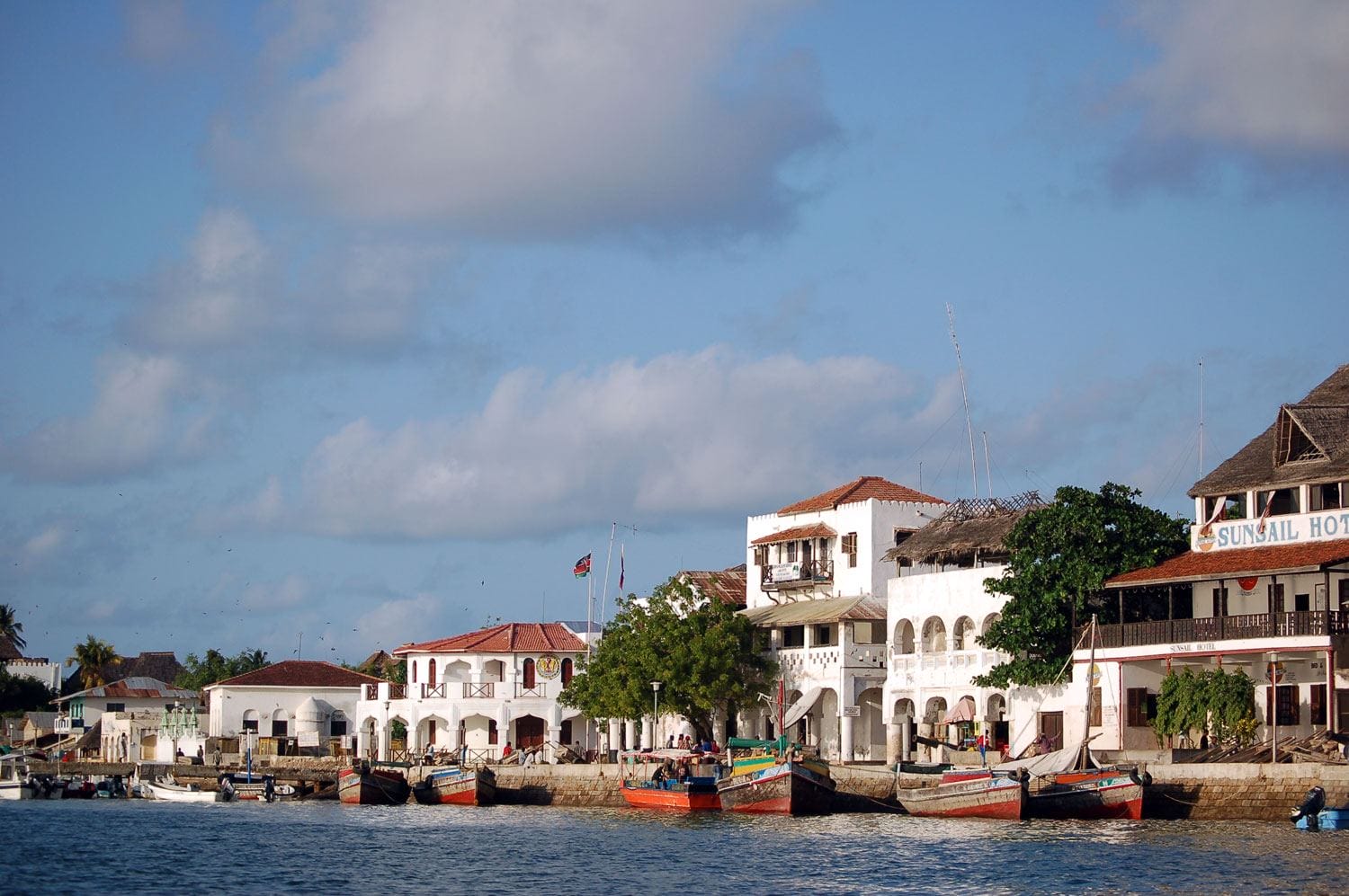 Colorful boats docked along a scenic waterfront with historic buildings under a partly cloudy sky.