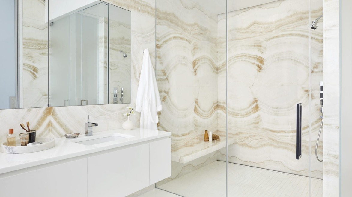 Modern bathroom with glass shower, elegant white sink, large mirrors, and beige marble walls featuring unique wave patterns.