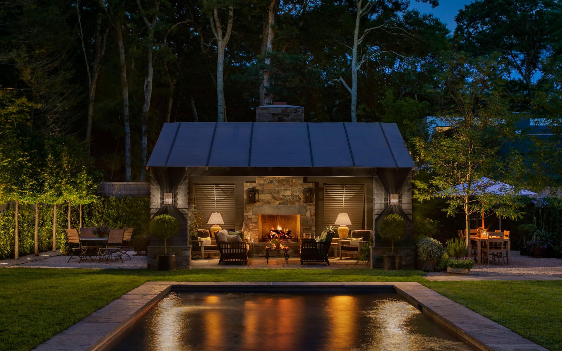 Outdoor living space with a lit fireplace and seating area, surrounded by trees, overlooking a serene pool at dusk.