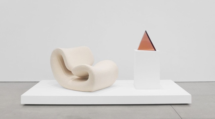 Curved white modern chair beside a geometric sculpture on a pedestal in a minimalist gallery setting