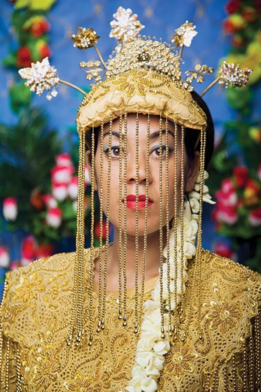 Woman in traditional gold attire and headpiece with floral accents, standing before a colorful background.