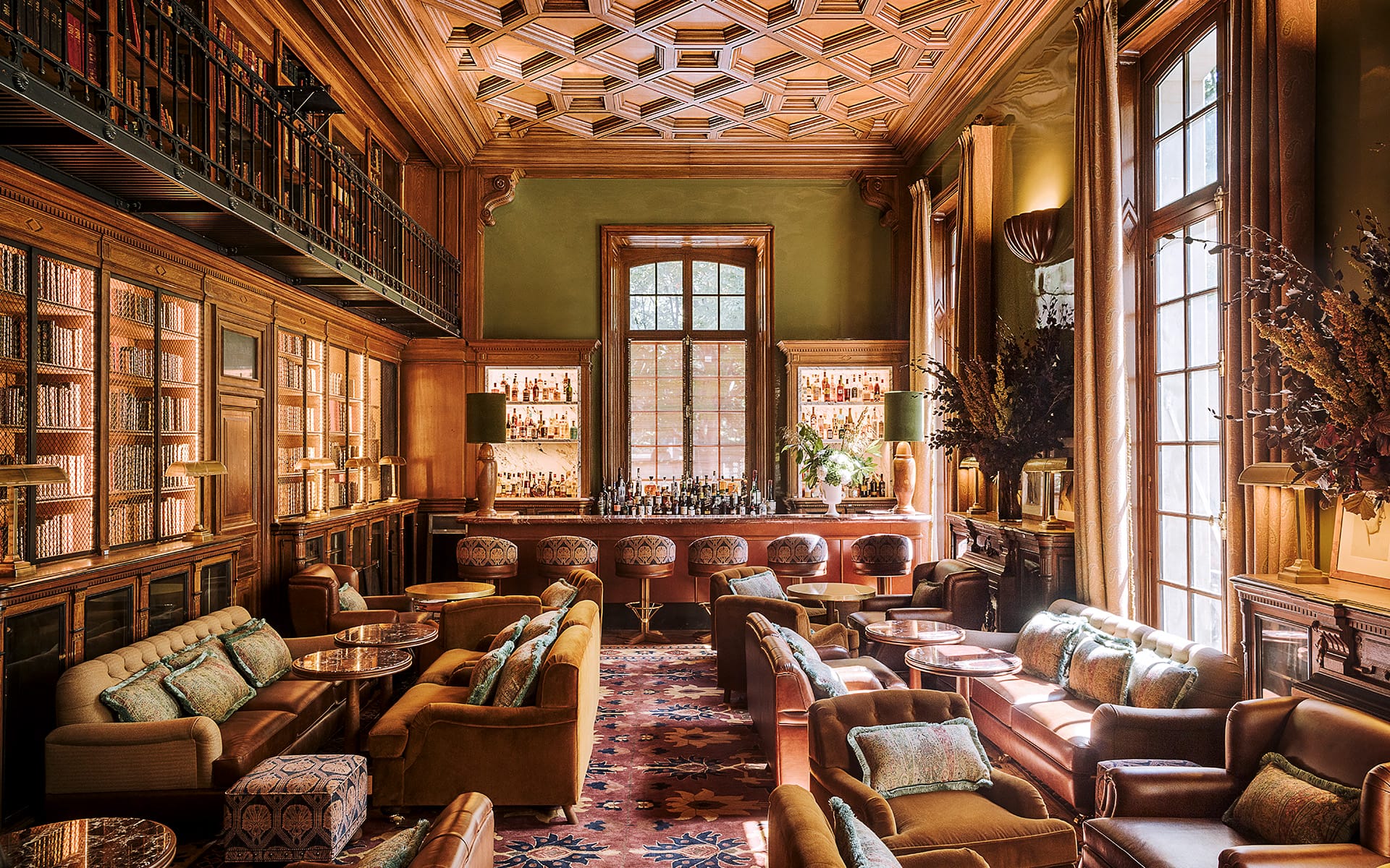 Elegant lounge with plush seating, wooden bookshelves, large windows, and a stocked bar against a warm-toned ceiling.