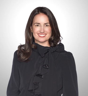 Person smiling, wearing a black blouse with ruffle details, against a light gray background.