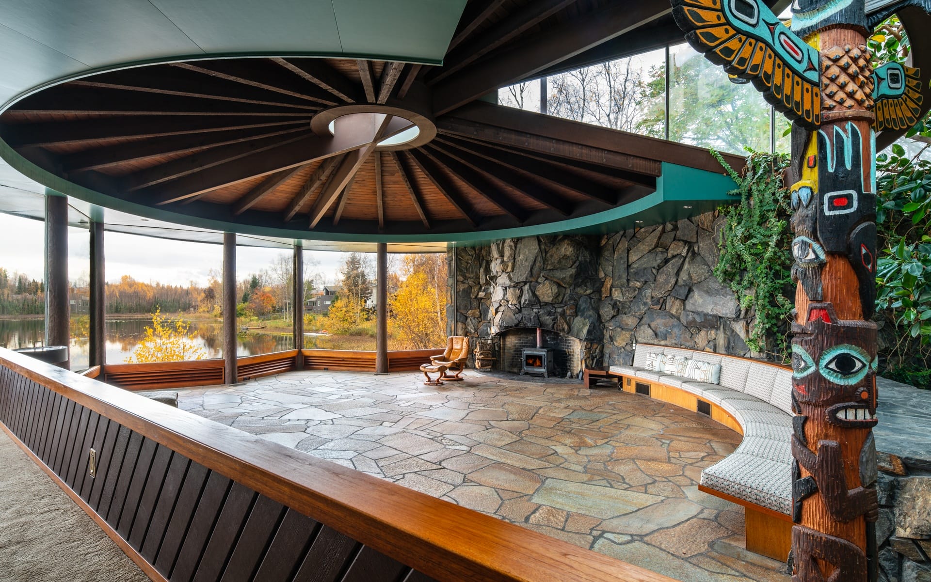 Circular room with large windows, stone floor, fireplace, and totem pole, overlooking autumn landscape and lake.