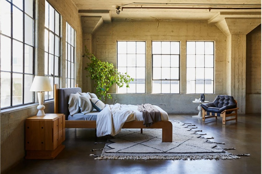 Spacious loft bedroom with large windows, wooden bed, cozy armchair, leafy plant, and modern decor.