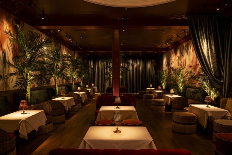 Luxurious dimly lit restaurant with elegant table settings, surrounded by tall plants and curtains, creating an intimate atmosphere.