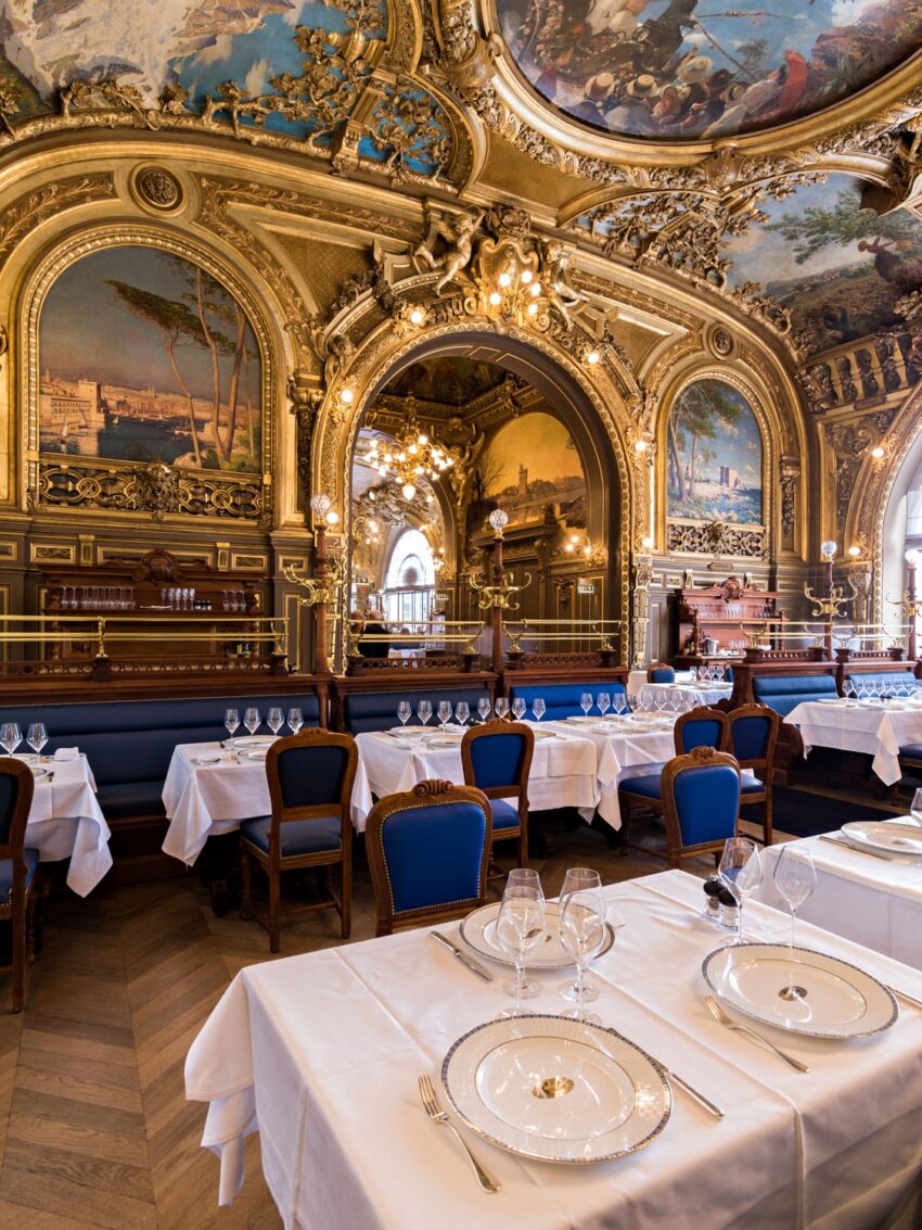Elegant dining room with ornate ceiling, chandeliers, and tables set with white tablecloths and fine china.