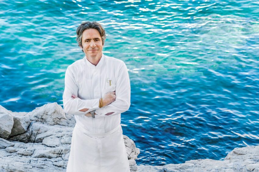 Chef standing on rocks by the sea with arms crossed, wearing a white uniform, blue water in the background.