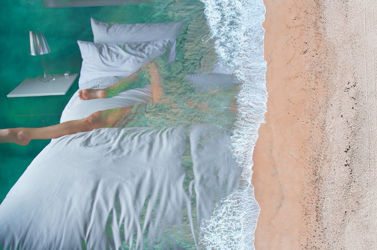 Surreal image of a bed merging with an ocean shore, depicting waves washing over white bedding next to sandy beach.