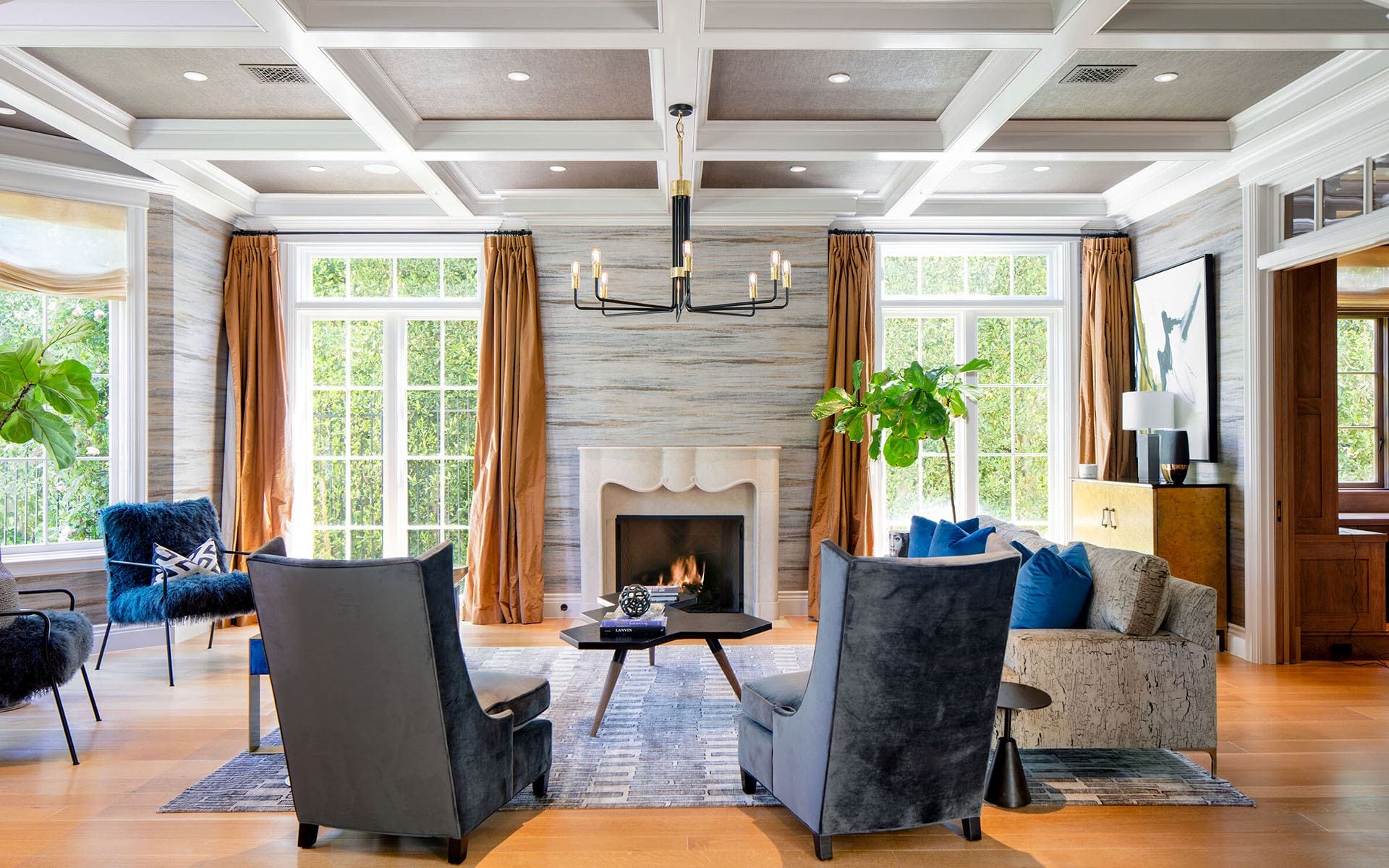 Elegant living room with coffered ceiling, fireplace, velvet chairs, and large windows with gold curtains.