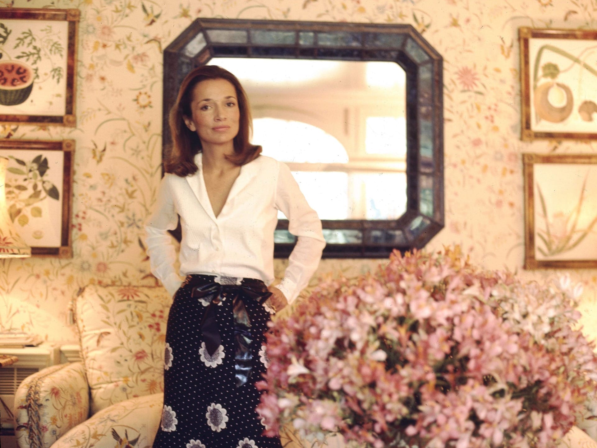 Person standing in a floral-themed room with a large mirror in the background and a bouquet of pink flowers in the foreground.