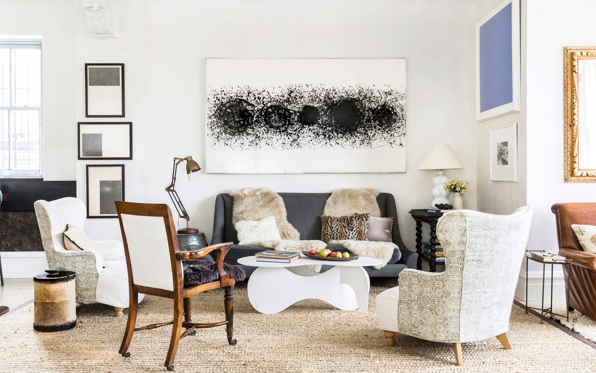 Modern living room with eclectic decor, featuring a gray sofa, abstract art, patterned chairs, and a unique coffee table.