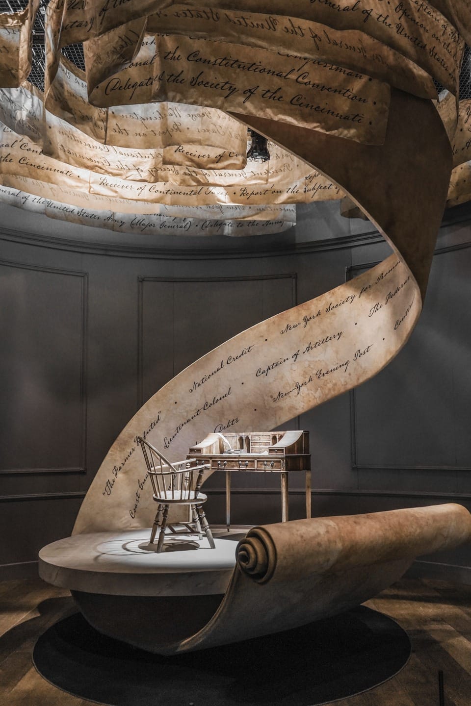 Spiral scroll sculpture with historical text, chair, and desk in dimly lit room, emphasizing themes of history and governance.