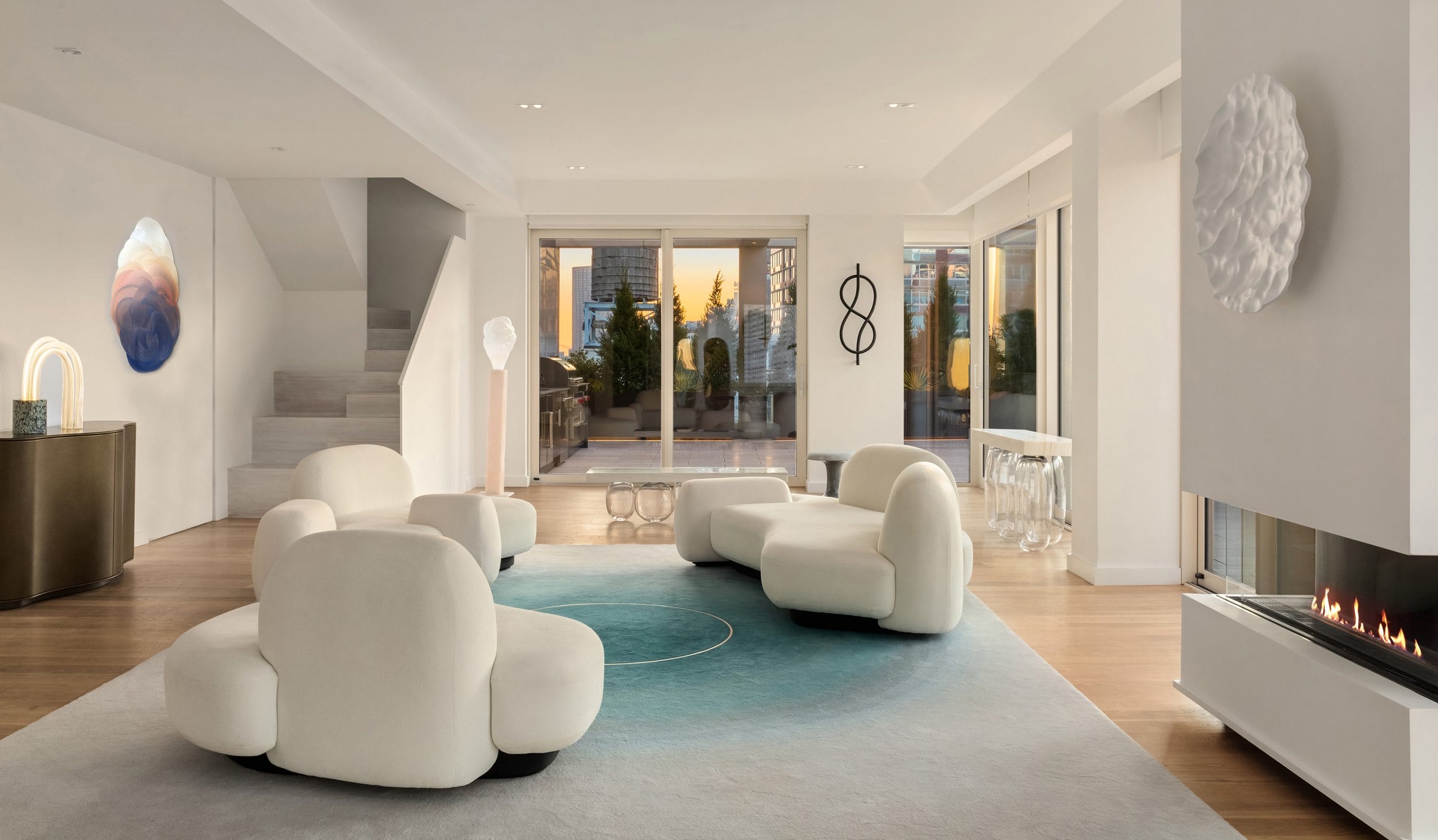 Modern living room with unique white chairs, abstract art on walls, blue gradient rug, fireplace, and large windows showing sunset.