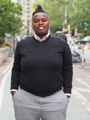 Person wearing a black sweater and gray pants stands on a city street with hands in pockets, greenery in the background.