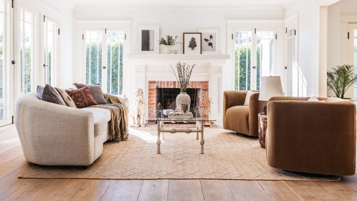 Bright living room with modern furniture, large windows, and a fireplace, featuring a cozy rug and decorative plants.