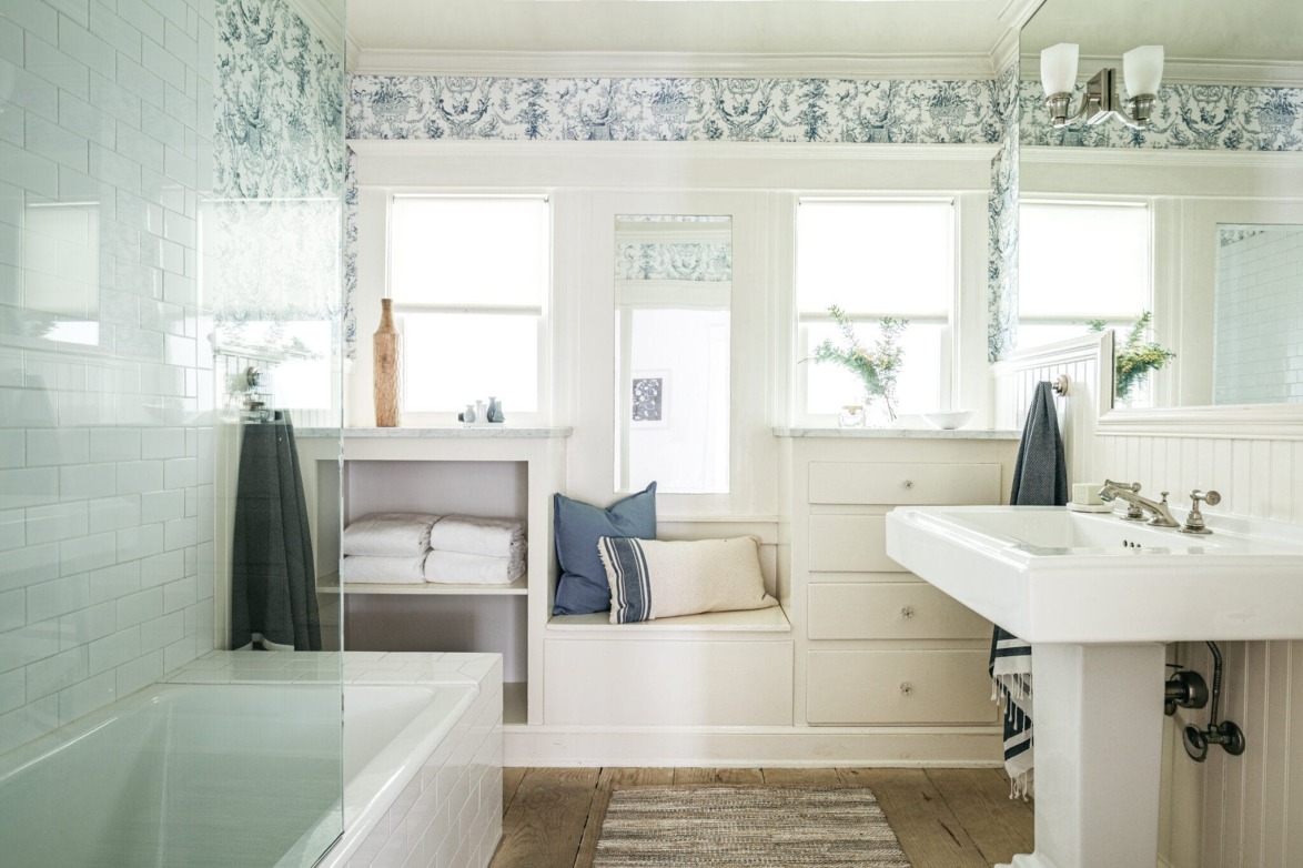 Bright bathroom with floral wallpaper, pedestal sink, bench seat, and window shelves holding towels and decor items.