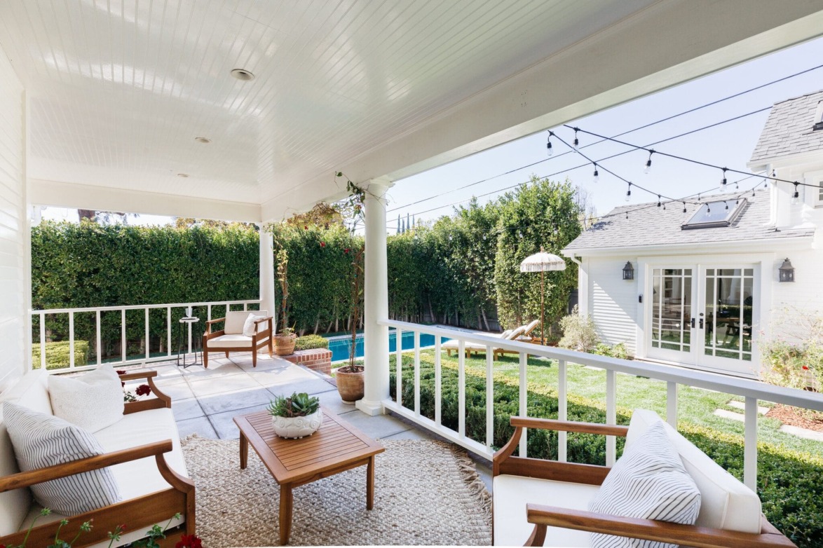 Covered patio with white furniture overlooking a lush backyard, pool, and string lights on a sunny day.