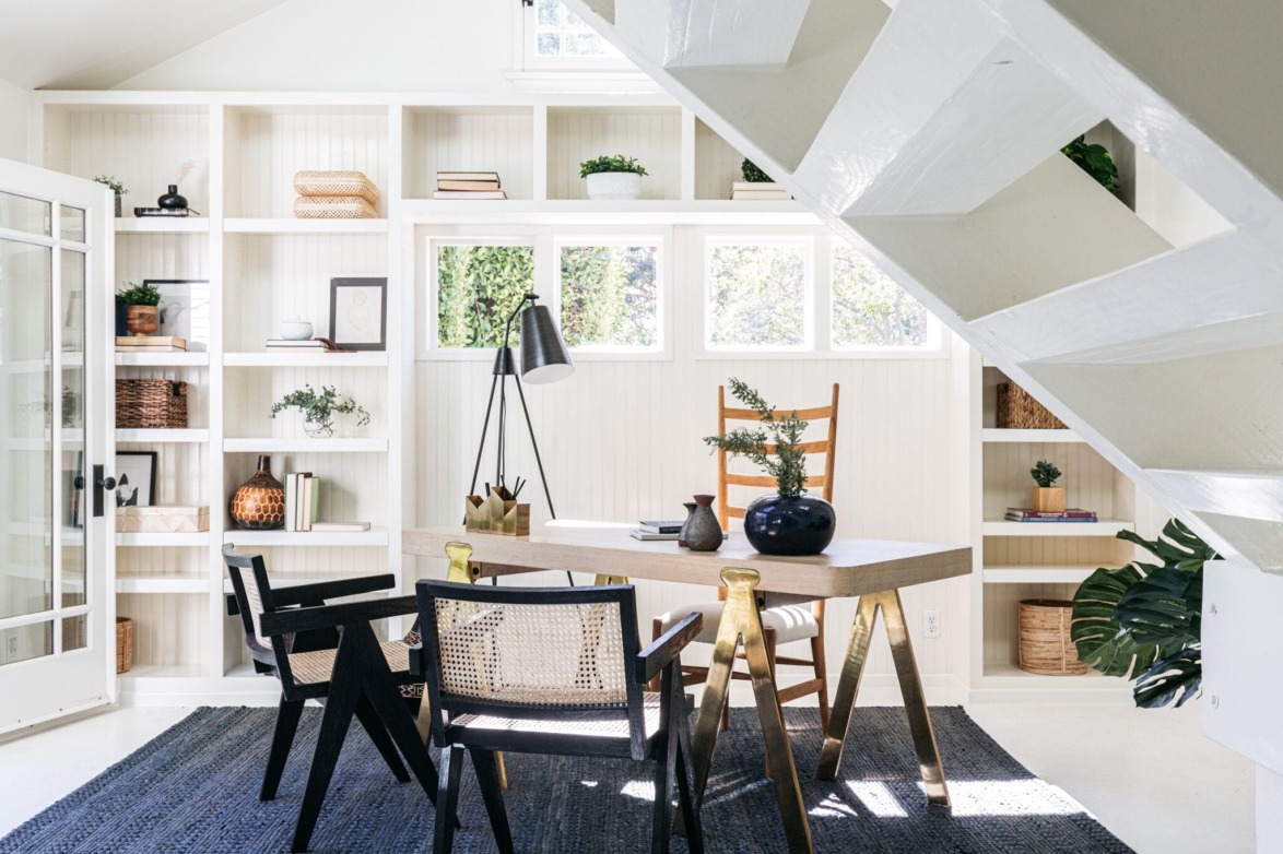 Modern home office with wooden desk, black chairs, and built-in shelves under a white staircase. Cozy and stylish decor.