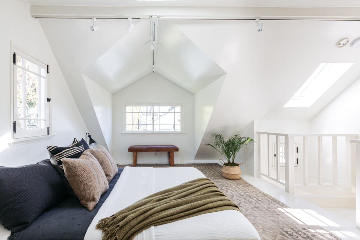Bright attic bedroom with large windows, a cozy bed with pillows, green throw blanket, potted plant, and natural light.