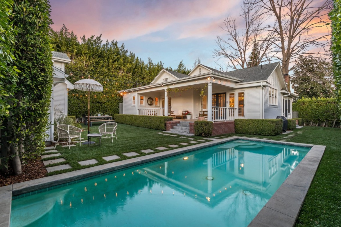 Charming white house with a porch and a pool in the backyard, surrounded by green hedges, under a colorful sunset sky.