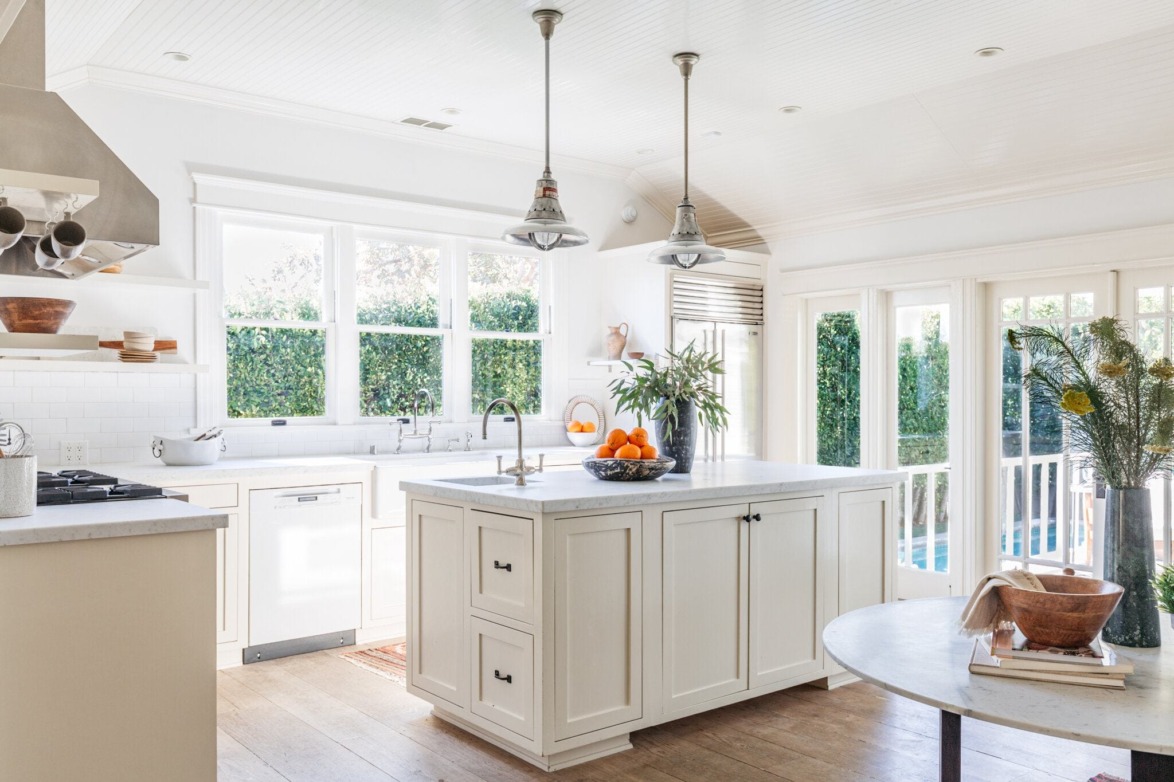 Bright, spacious kitchen with white cabinets, island with fruit bowl, large windows, and greenery outside.