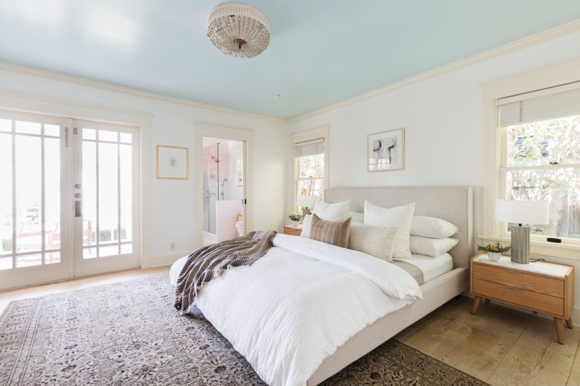 Modern bedroom with a cozy bed, soft white bedding, large windows, and elegant decor, featuring a stylish chandelier.