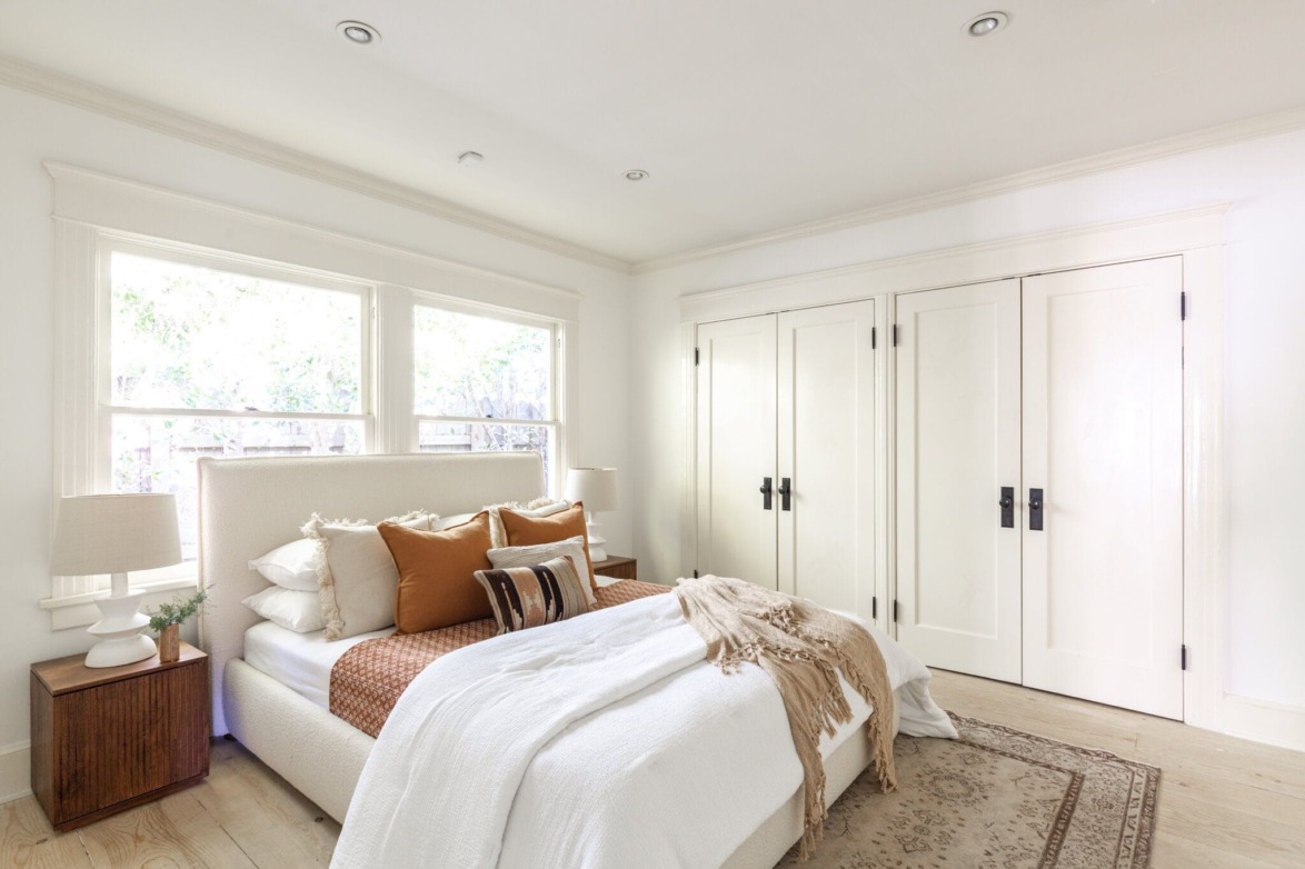 Bright bedroom with large windows, white bedding, and brown accent pillows; two lamps on side tables; double door closet.