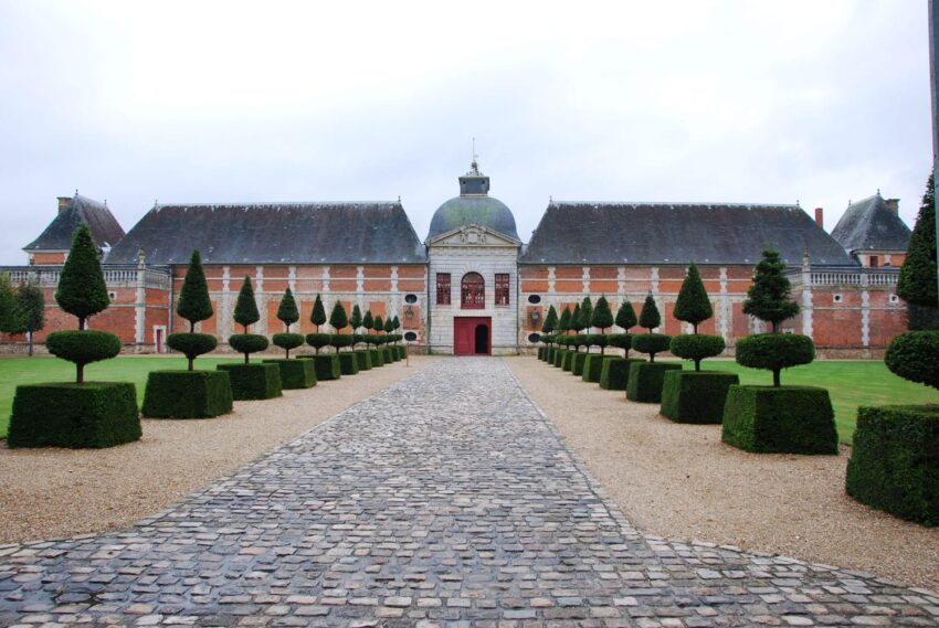 Historic brick building with arched entrance, surrounded by manicured trees and a cobblestone path on a cloudy day.