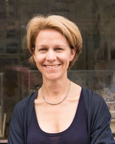 Smiling person with short hair in a dark top and necklace standing outdoors in front of a window display.