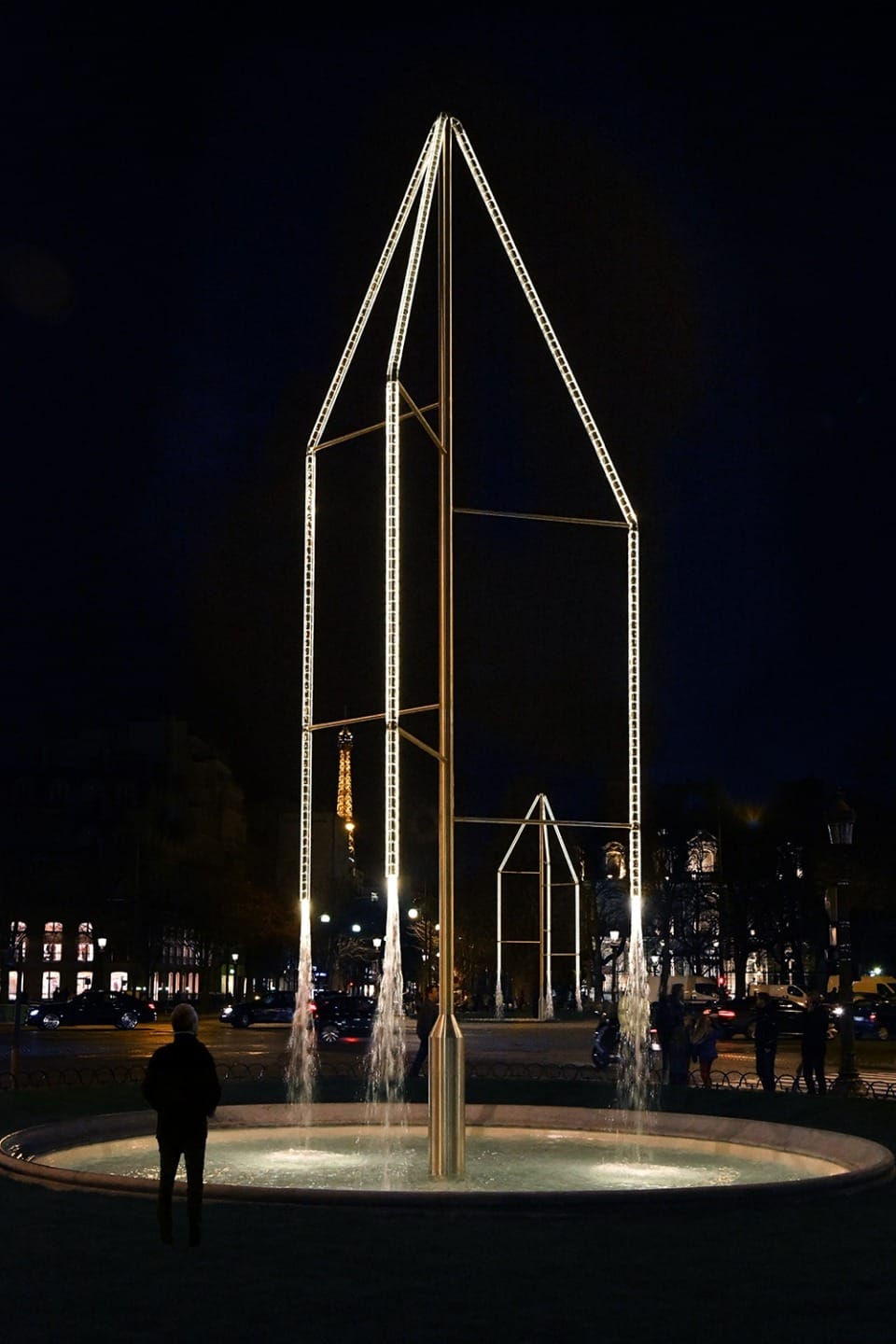Illuminated geometric sculpture with fountains in a city square at night, featuring a tall, glowing framework structure.