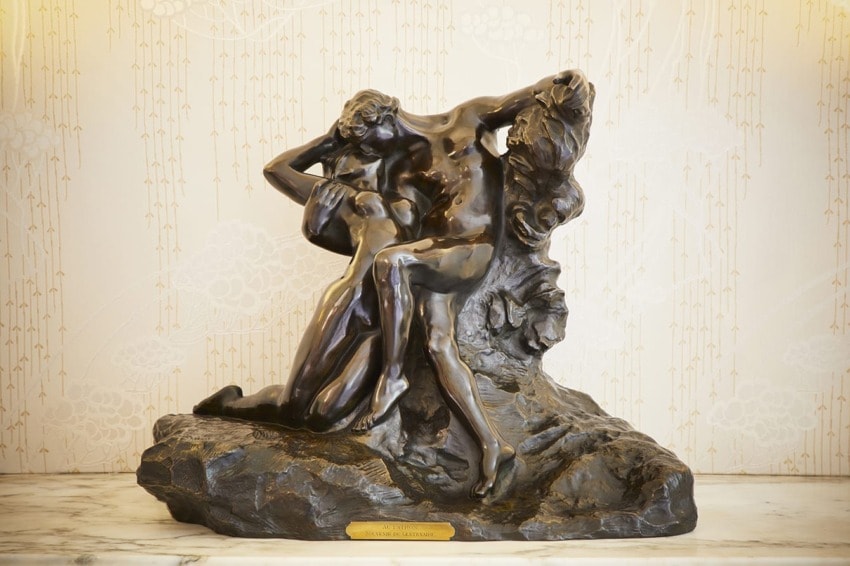 Bronze sculpture of a seated woman and kneeling man, embracing passionately on a textured base, set against decorative wallpaper.