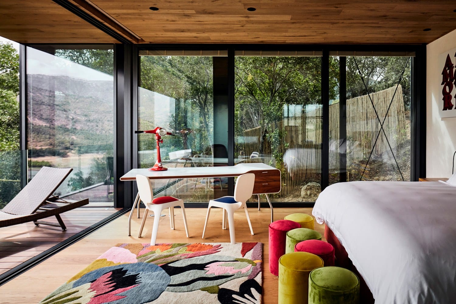 Modern bedroom with large glass windows, a desk, chairs, colorful stools, and a view of nature outside.