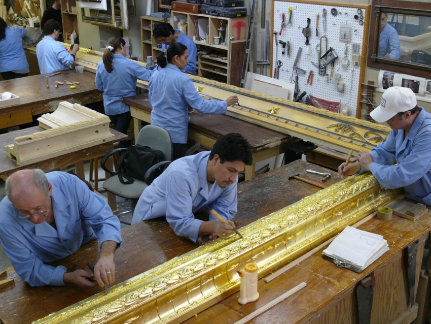 Artisans in blue coats work on ornate gold frames in a workshop with tools on the wall and wooden tables.