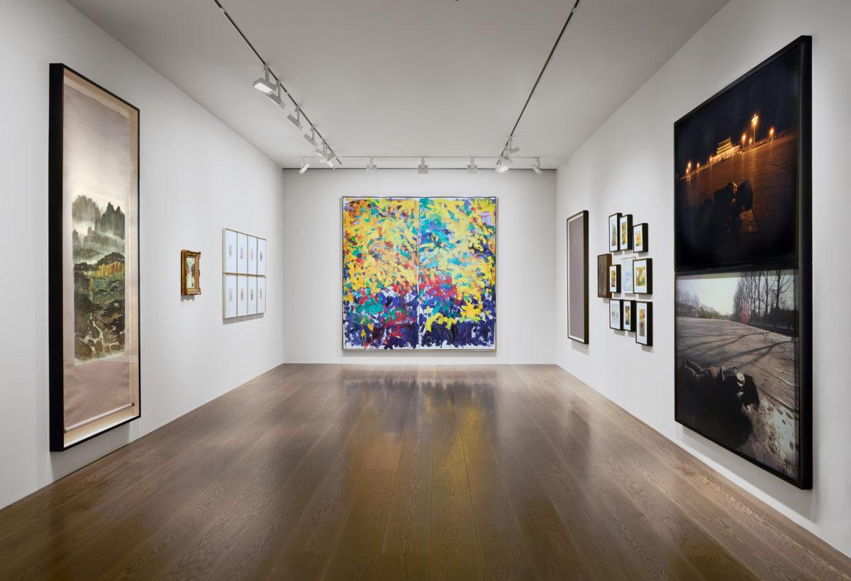 Contemporary art gallery interior with paintings and photographs displayed on white walls and wooden floor.