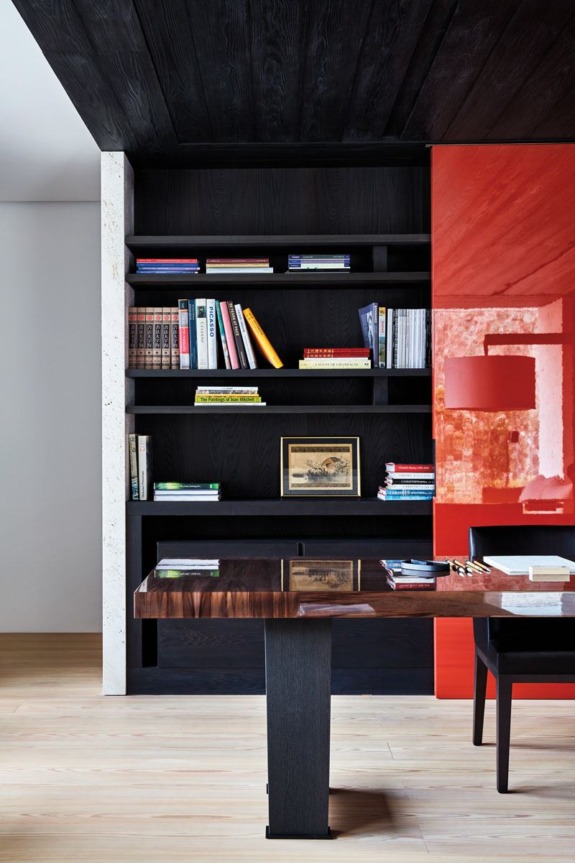 Modern office with black shelves, books, and a painting, red accent wall, wooden desk, and a chair on light wood flooring.