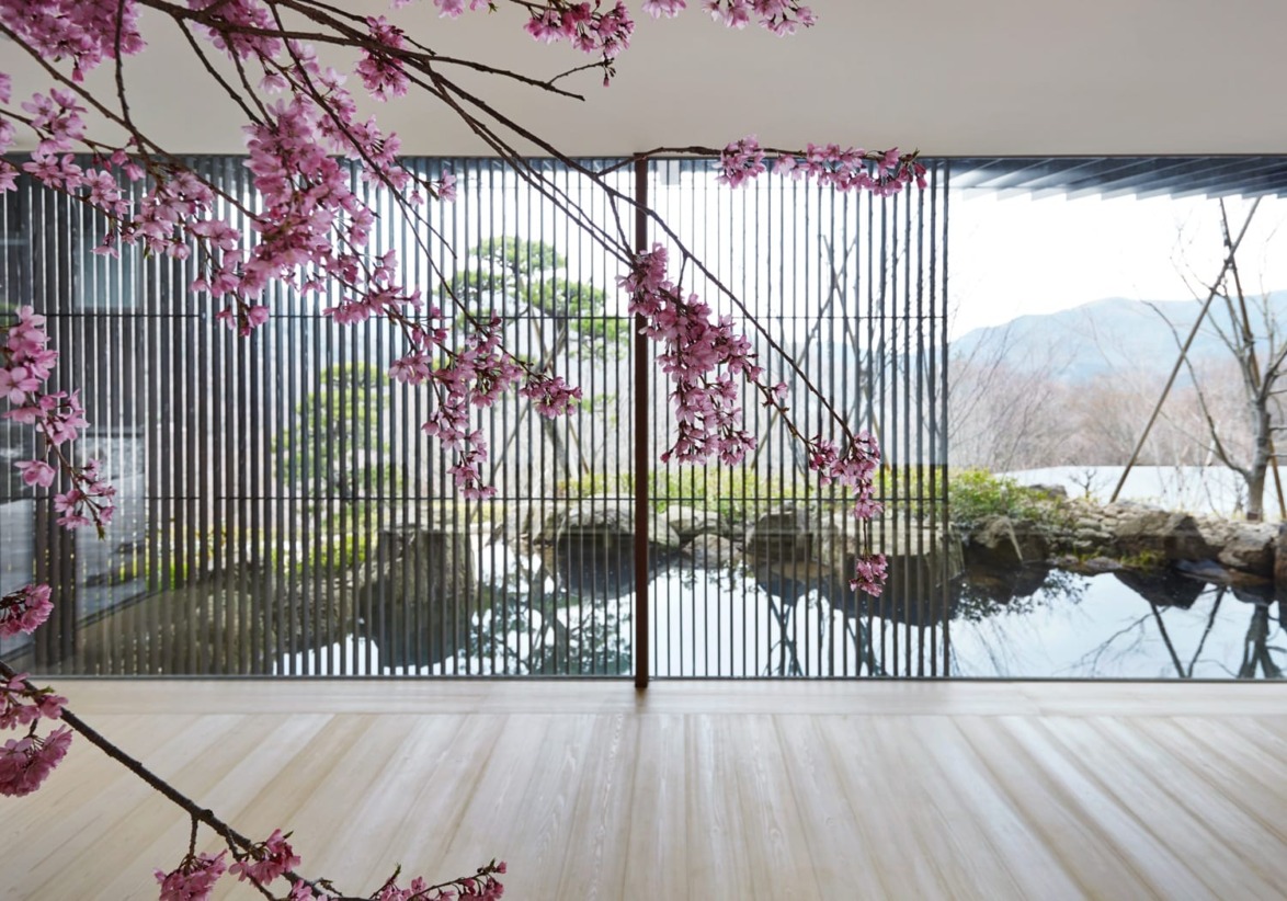 Cherry blossoms near a modern glass building with a serene mountain and water background visible through large windows.