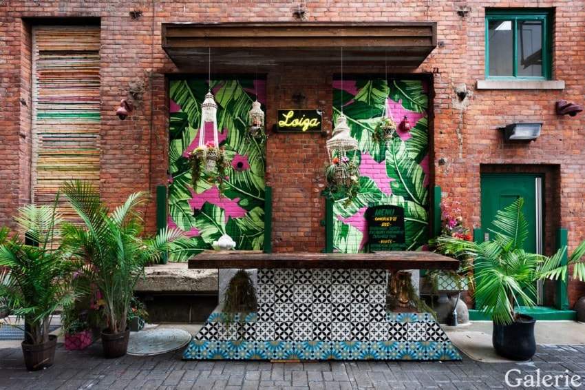 Colorful patio with tropical mural, potted plants, and a patterned bar, set against a red brick wall with a green door.