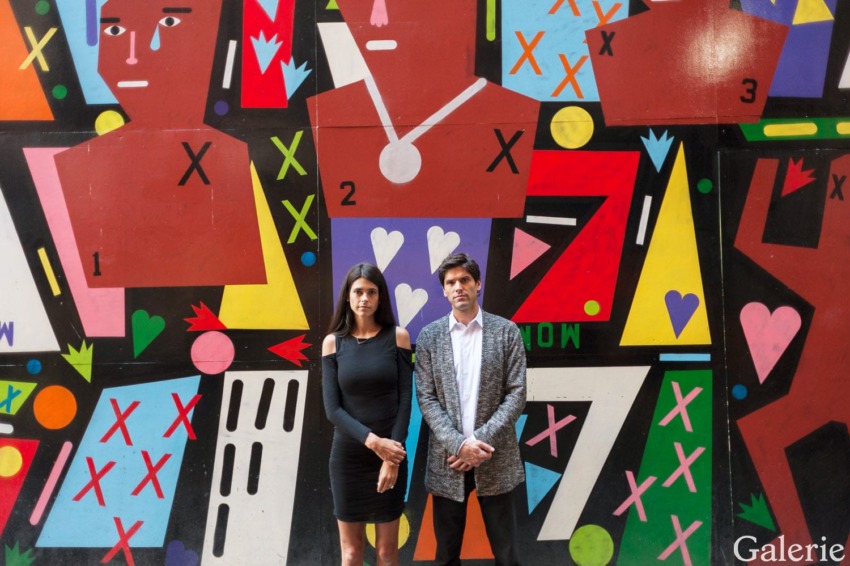 Two people standing in front of a colorful geometric mural with stylized figures and abstract shapes.