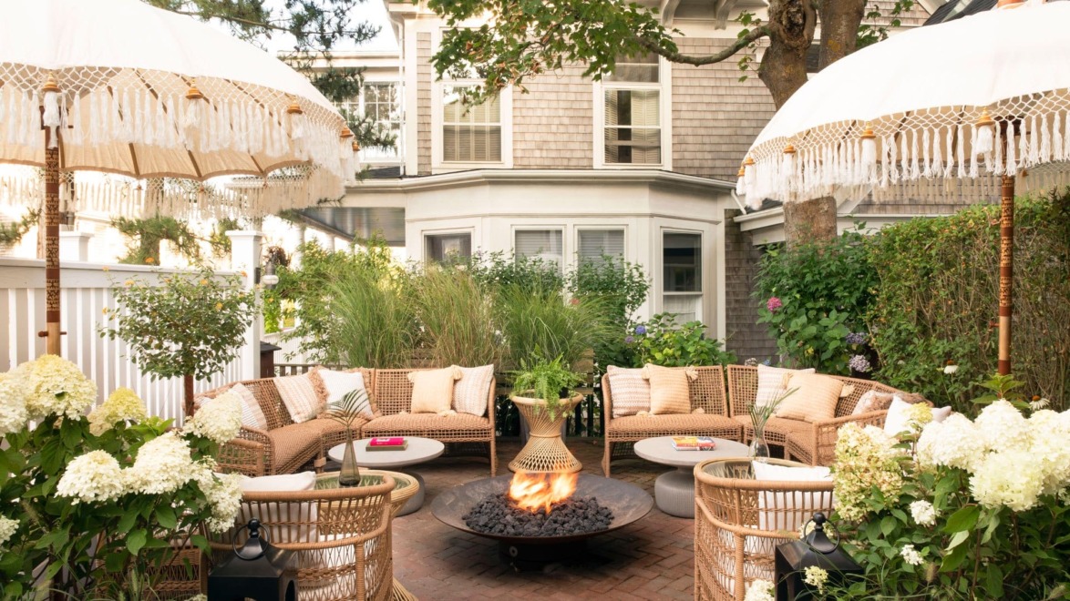 Outdoor patio with wicker furniture, white cushions, umbrellas, fire pit, and lush greenery in a cozy backyard setting.