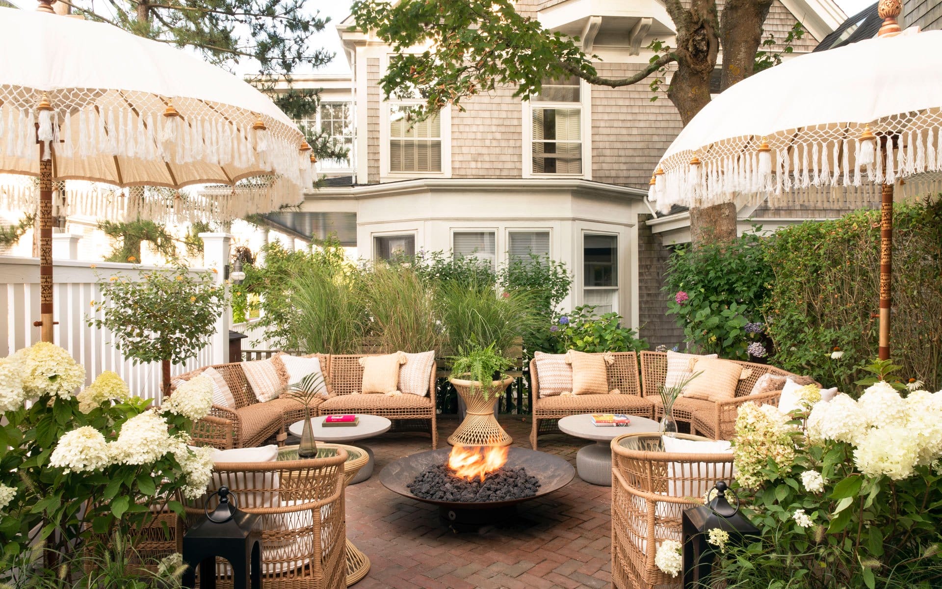 Outdoor patio with wicker furniture, white cushions, umbrellas, fire pit, and lush greenery in a cozy backyard setting.