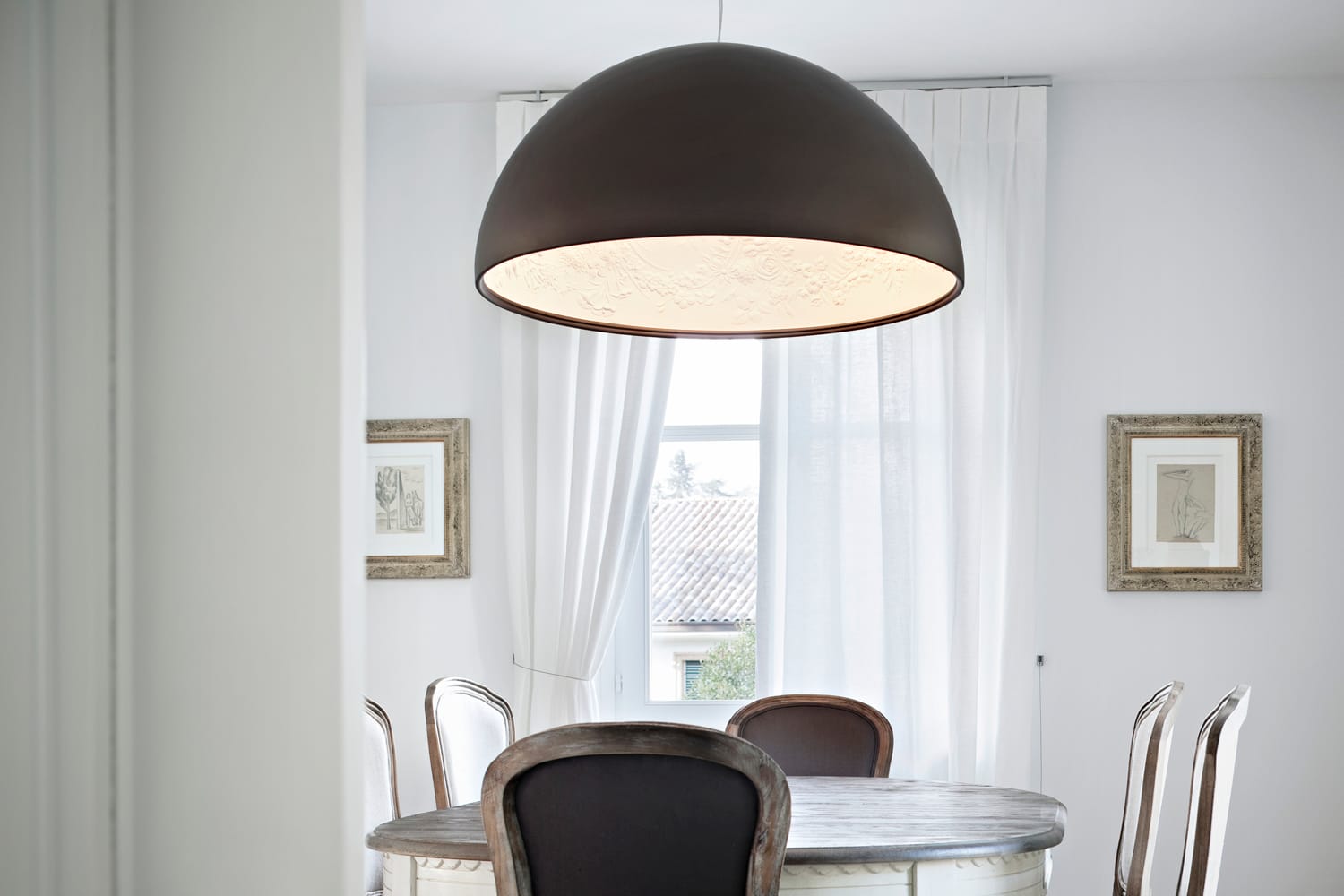 Modern dining room with a large pendant light above a round table and framed artworks on white walls.