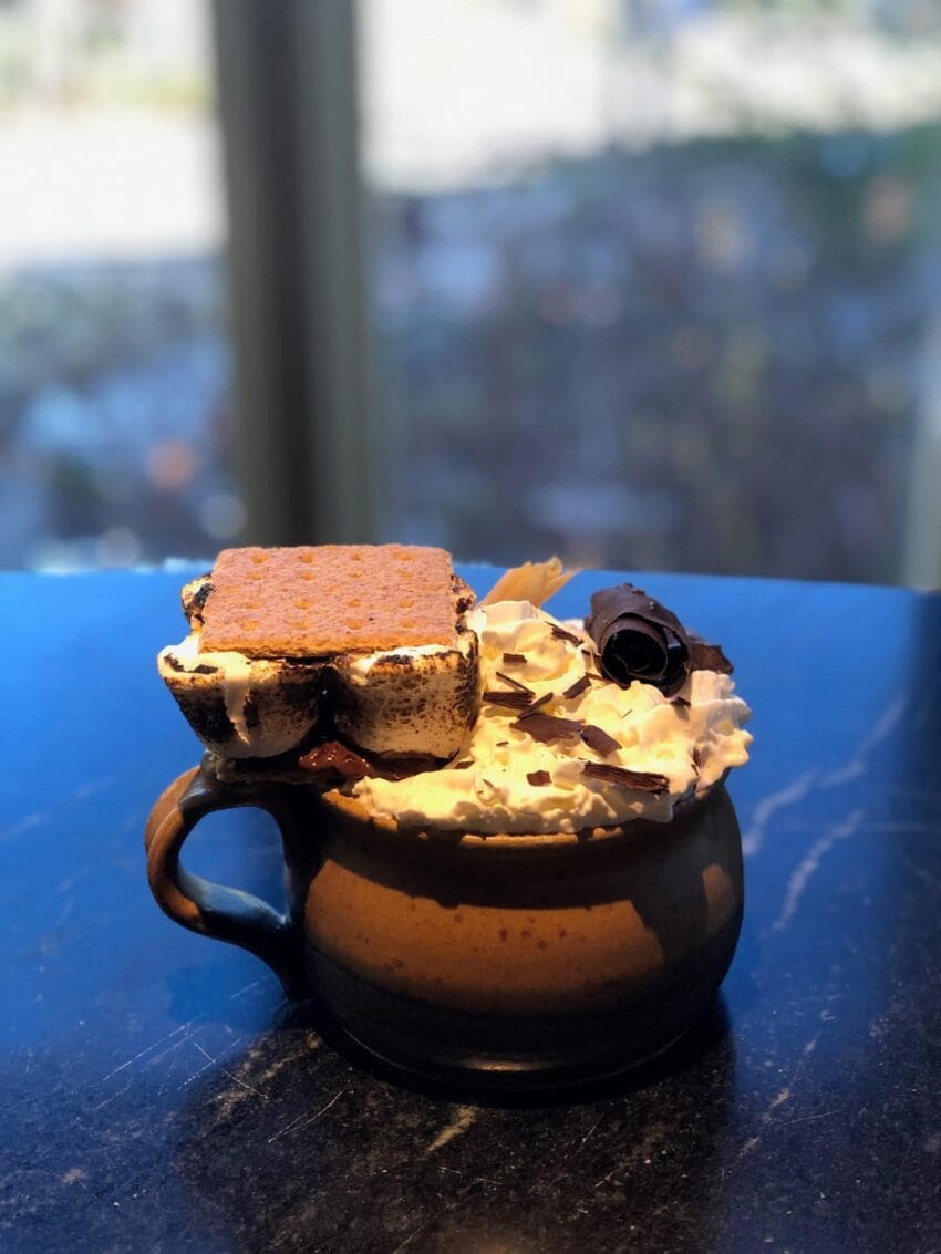 Decorative hot chocolate topped with whipped cream, chocolate curls, and a graham cracker marshmallow square on a table.