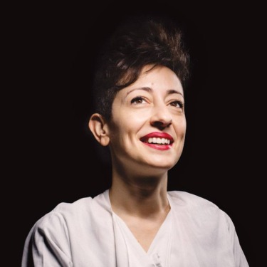 Person smiling brightly under a spotlight, wearing a white shirt against a dark background.