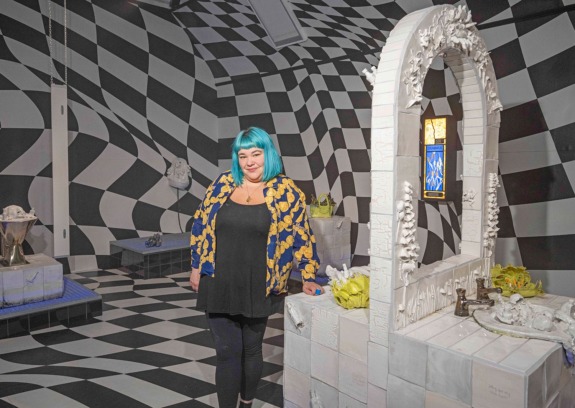A person with blue hair stands in a room with a black and white checkered pattern, next to a white arch sculpture.