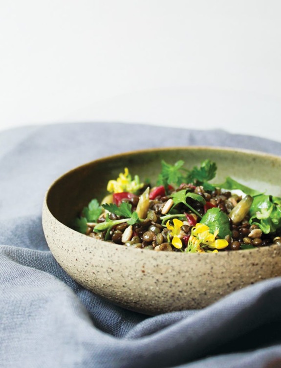 Bowl of lentil salad with seeds, herbs, and yellow flowers on a gray cloth background.